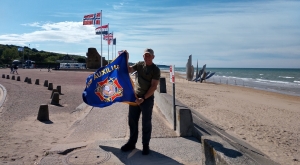 Jon Ring holds the VFW Auxiliary flag on Omaha Beach while touring Normandy for the 75th anniversary of D-Day.